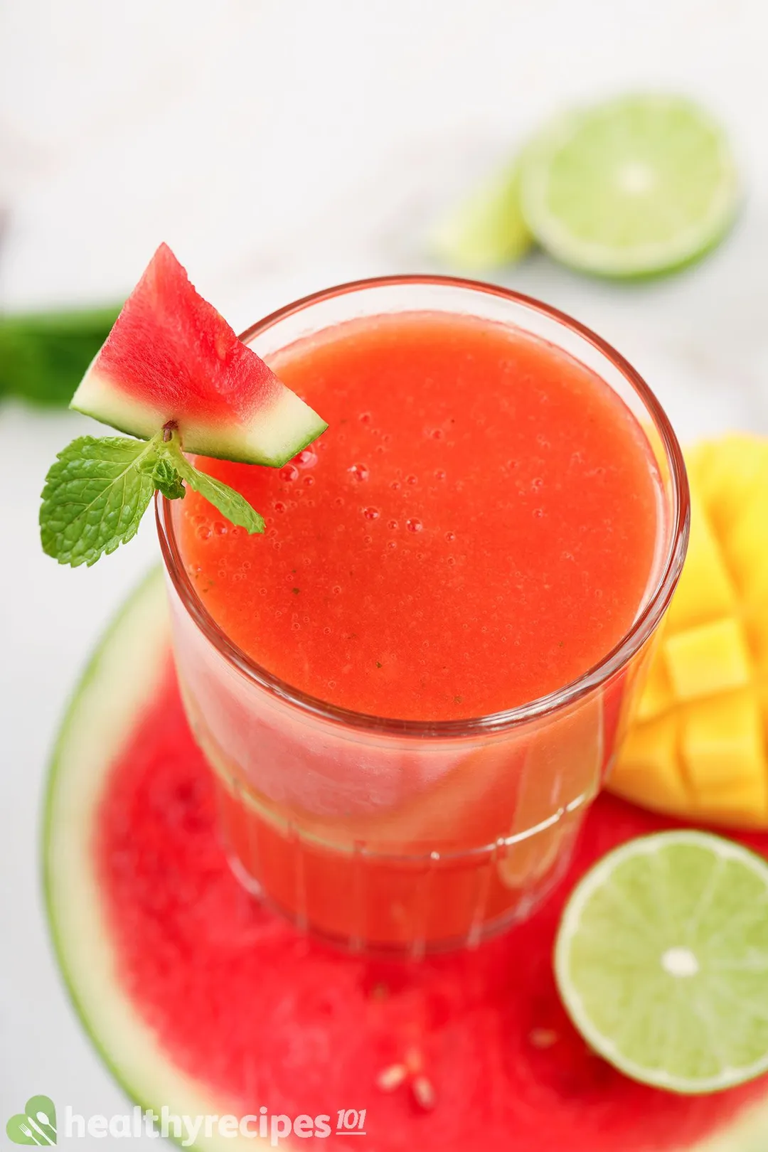 A high-angle shot of a Mango Watermelon Smoothie, half a lime, and a scored mango placed on half a watermelon.
