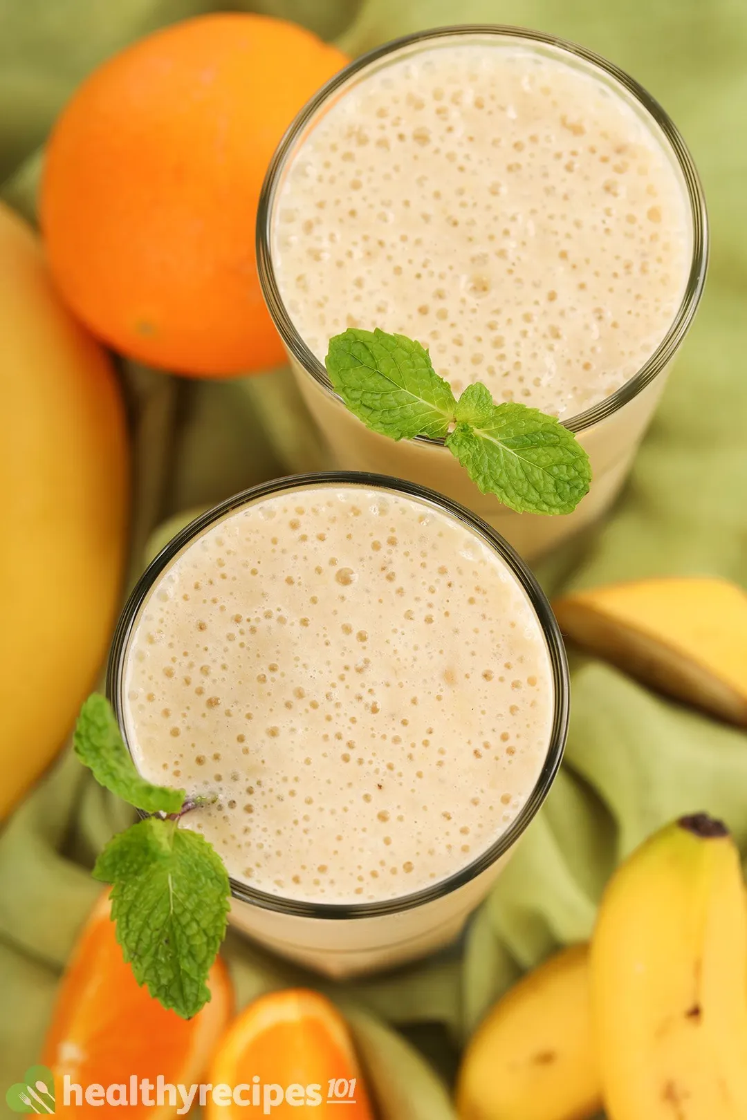 A high-angle shot of two glasses of Orange Banana Smoothie placed on a green cloth and near orange slices and unpeeled bananas.