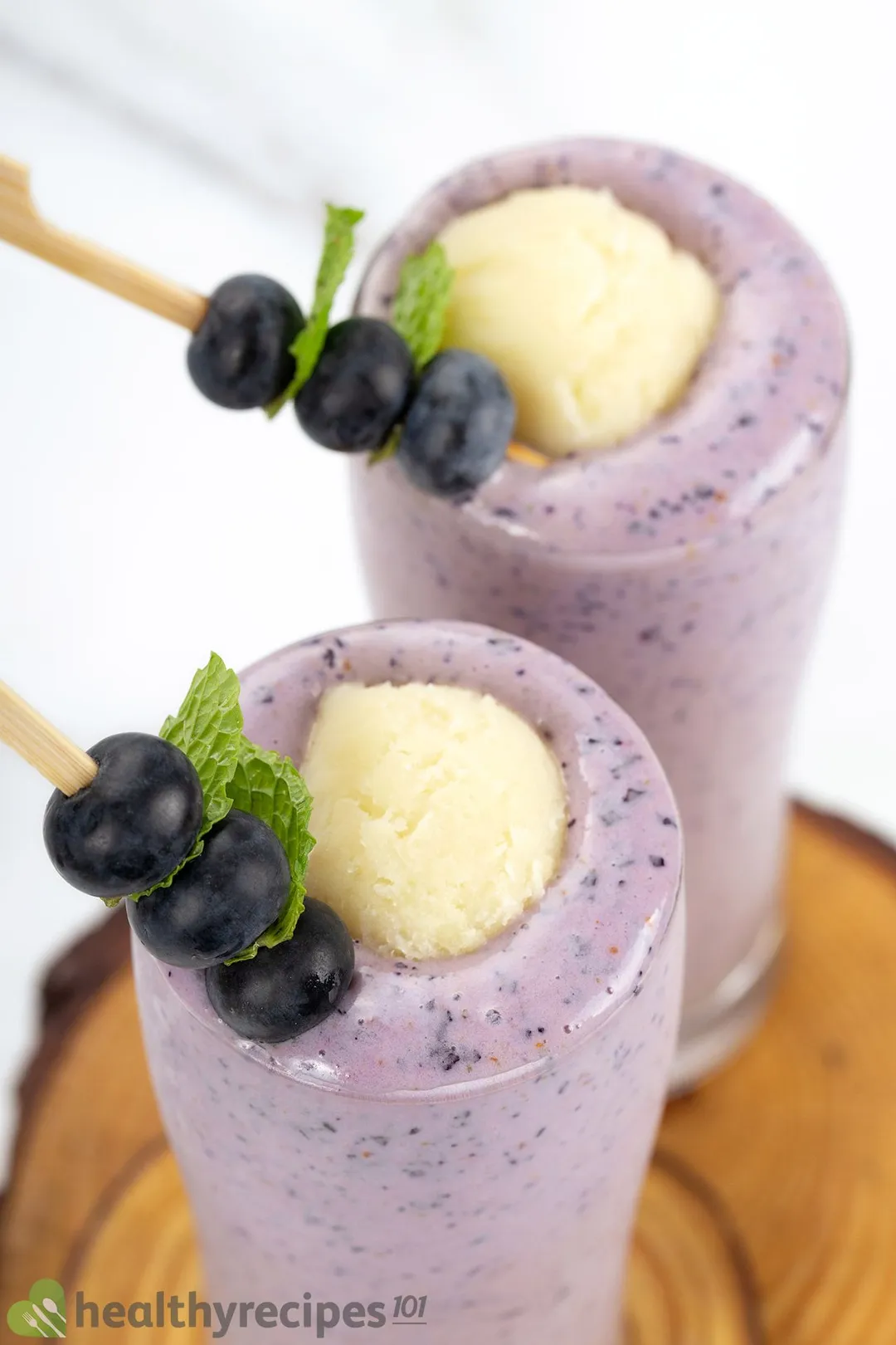 A high-angle shot of two tall blueberry banana smoothie glasses placed on wooden boards and garnished with blueberry skewers and ice cream scoops.
