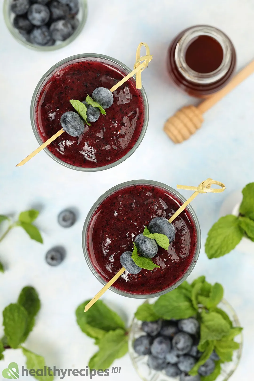 A high angle shot of two blueberry smoothie glasses, a small jar of honey, and a small plate of blueberries