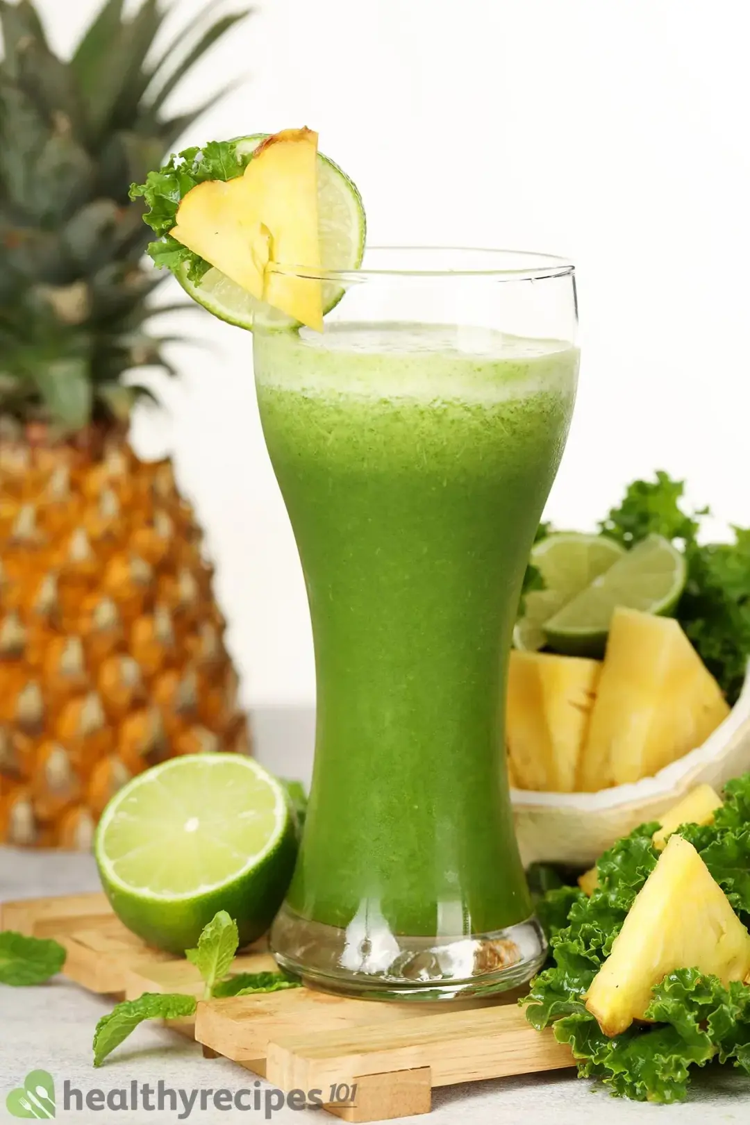 A glass of green mojito smoothie decorated with a slice of pineapple and lime surrounded by ingredients of the smoothie