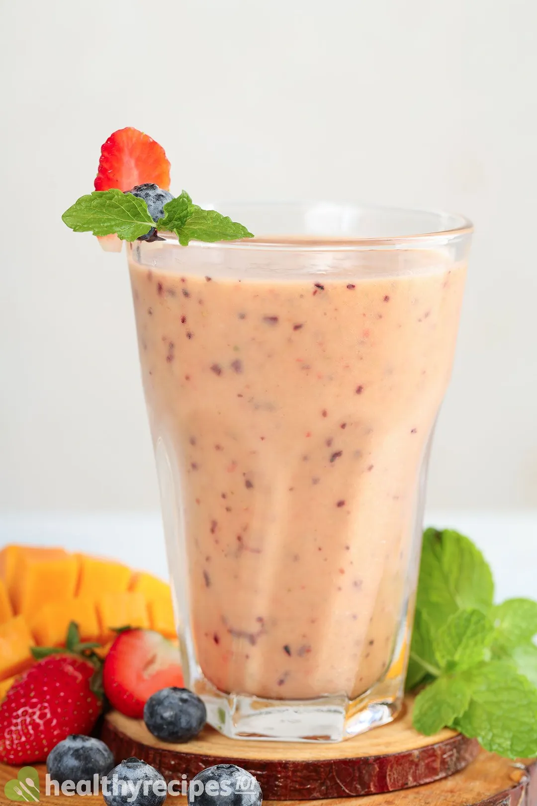 A glass of Mango Berry Smoothie placed on a wooden board near blueberries, strawberries, mint leaves, and a scored mango.