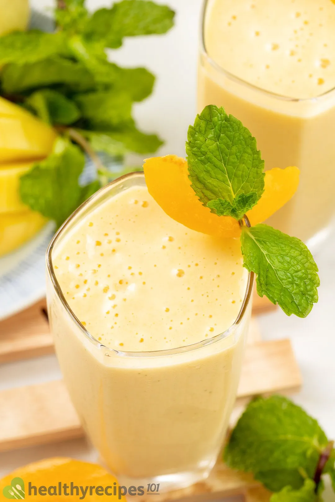A high-angle shot of two glasses of peach smoothie placed on a wooden board and near mint leaves.