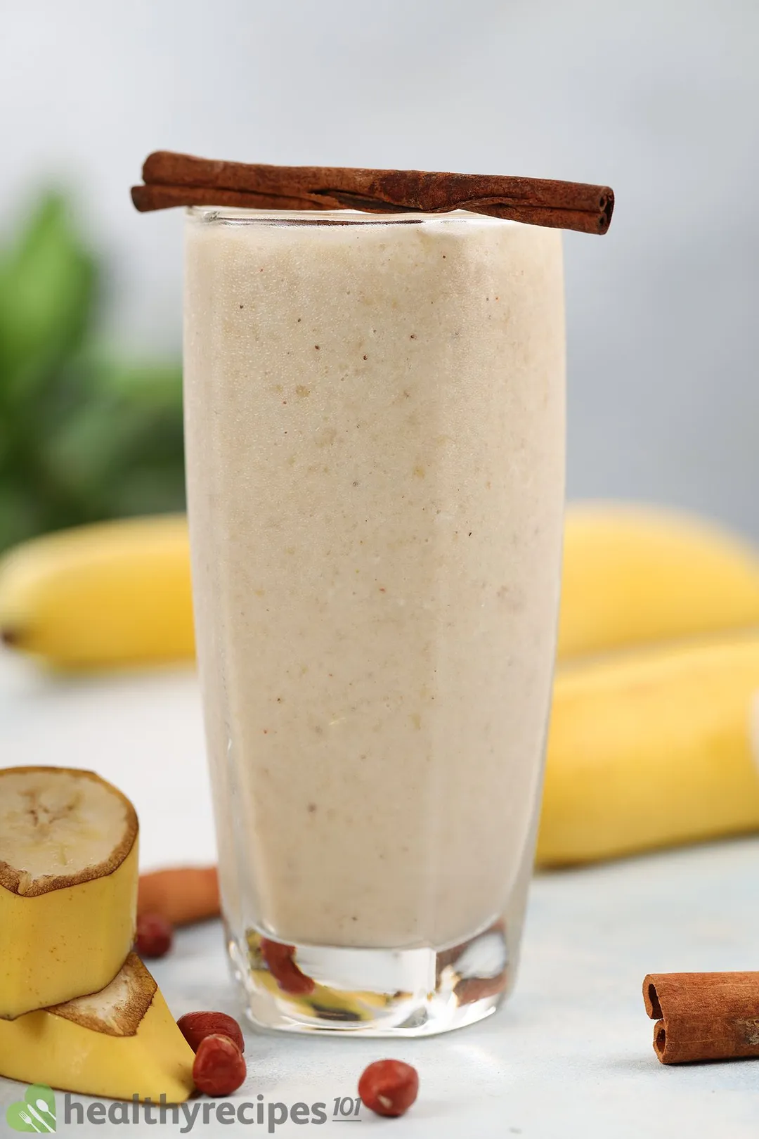 A glass of peanut butter banana smoothie surrounded by bananas, peanuts, and has a cinnamon stick placed on top of it.
