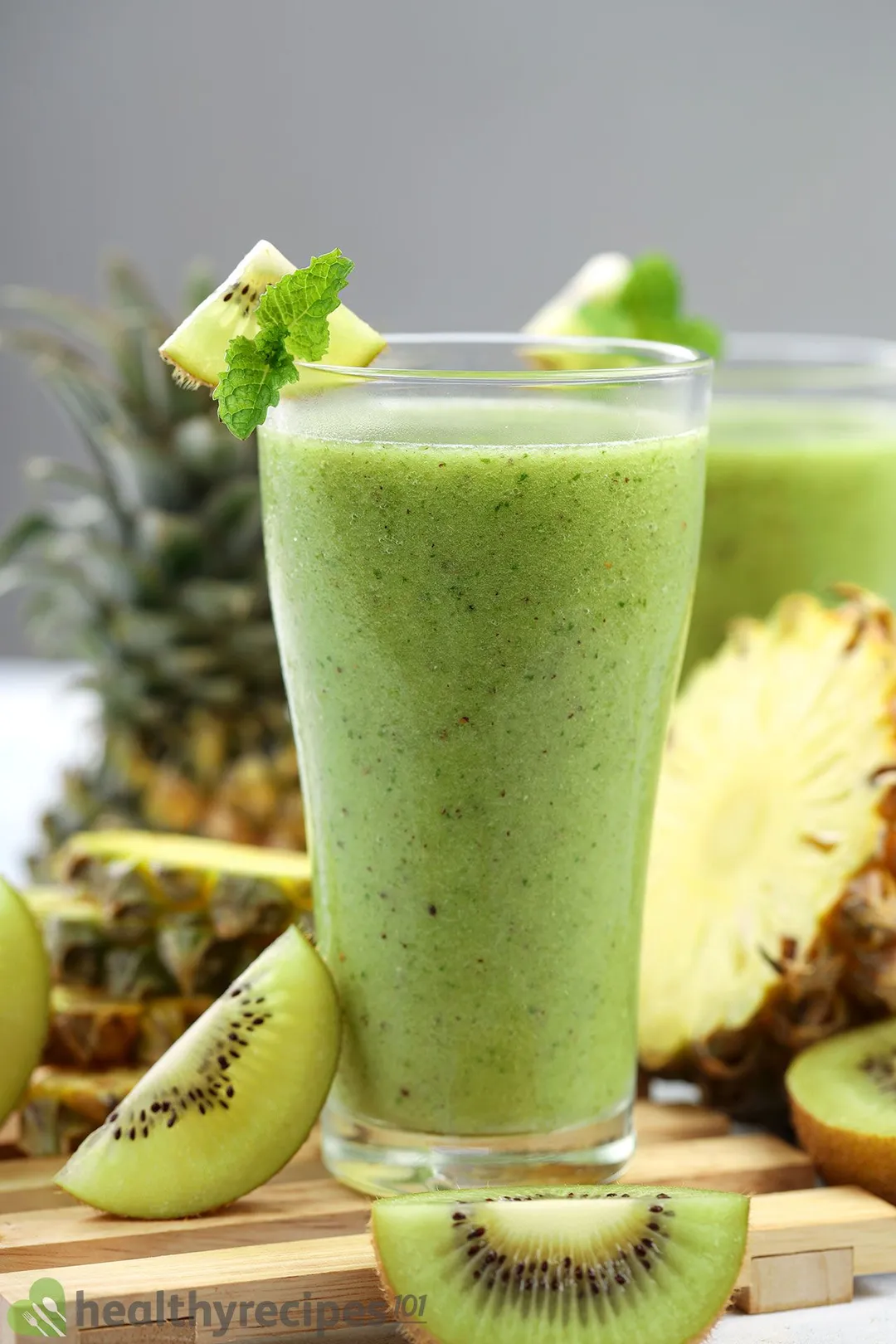 A glass of Pineapple Kiwi Smoothie placed on a wooden planks near green kiwi slices and pineapples.