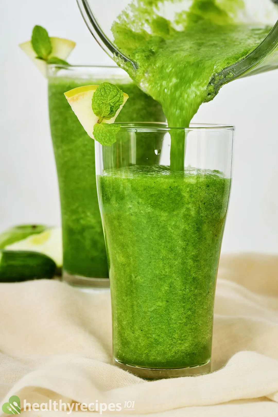 A glass jug pouring a vibrant green sour apple smoothie into a tall glass. In the back there's a similar glass.