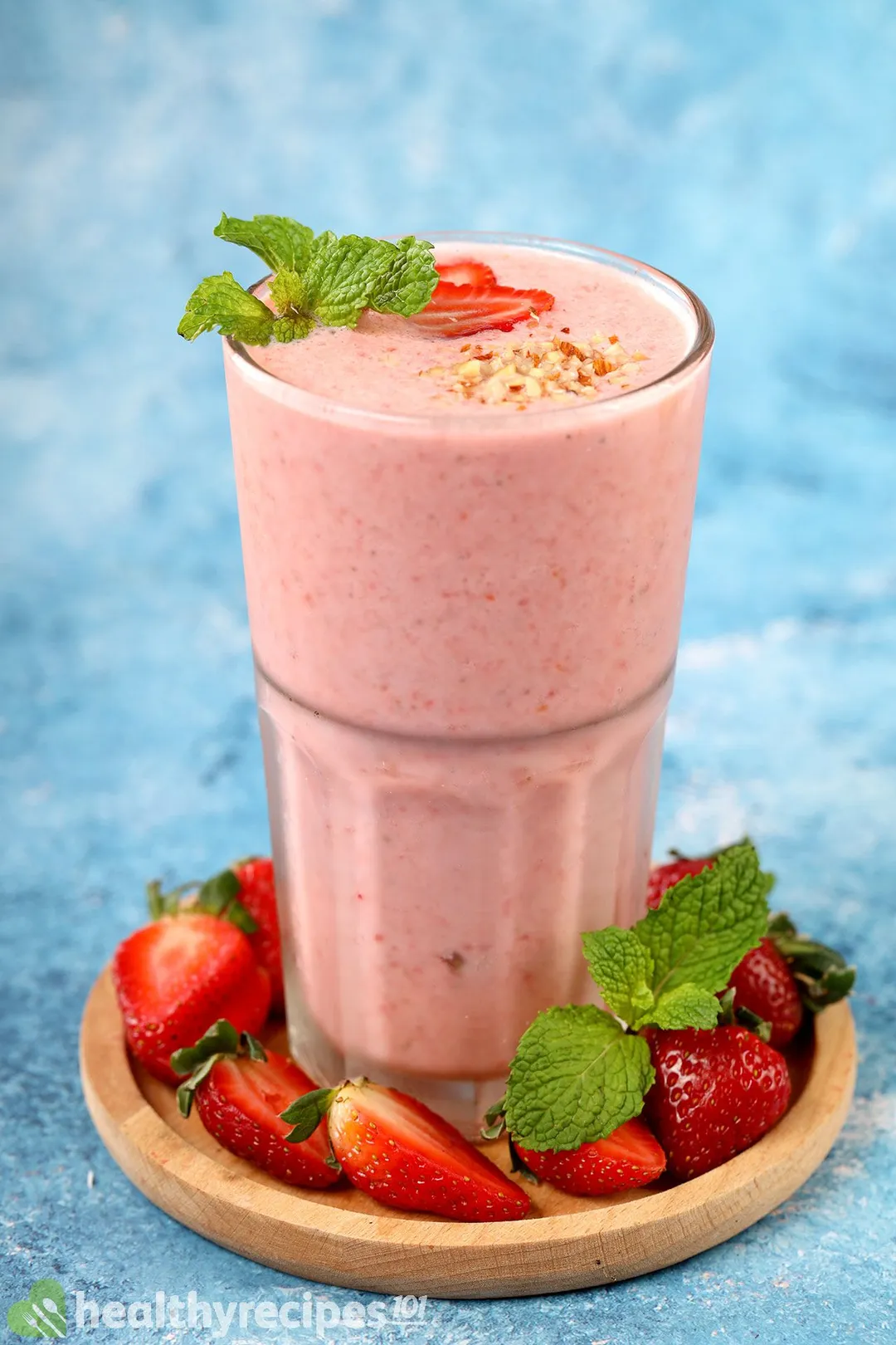 A glass of strawberry smoothie laid on a wooden coaster and surrounded by sliced strawberries and mint leaves.