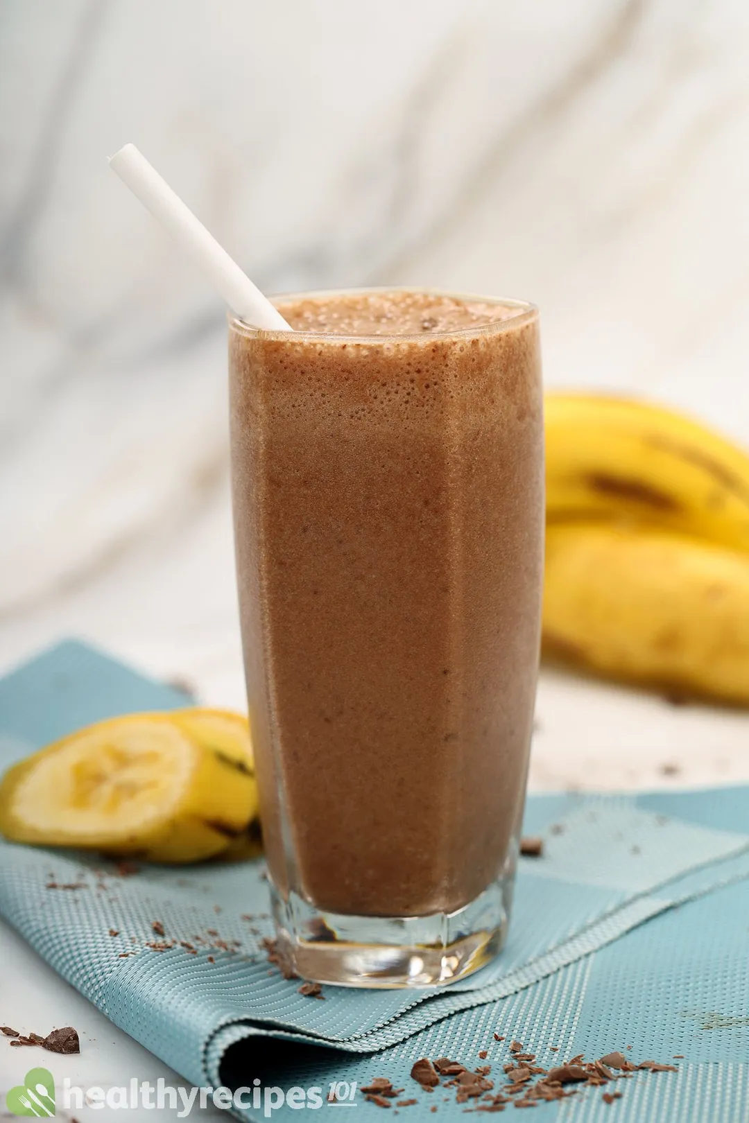 A tall glass of choolate banana smoothie placed on a blue cloth with unpeeled yellow bananas in the back.