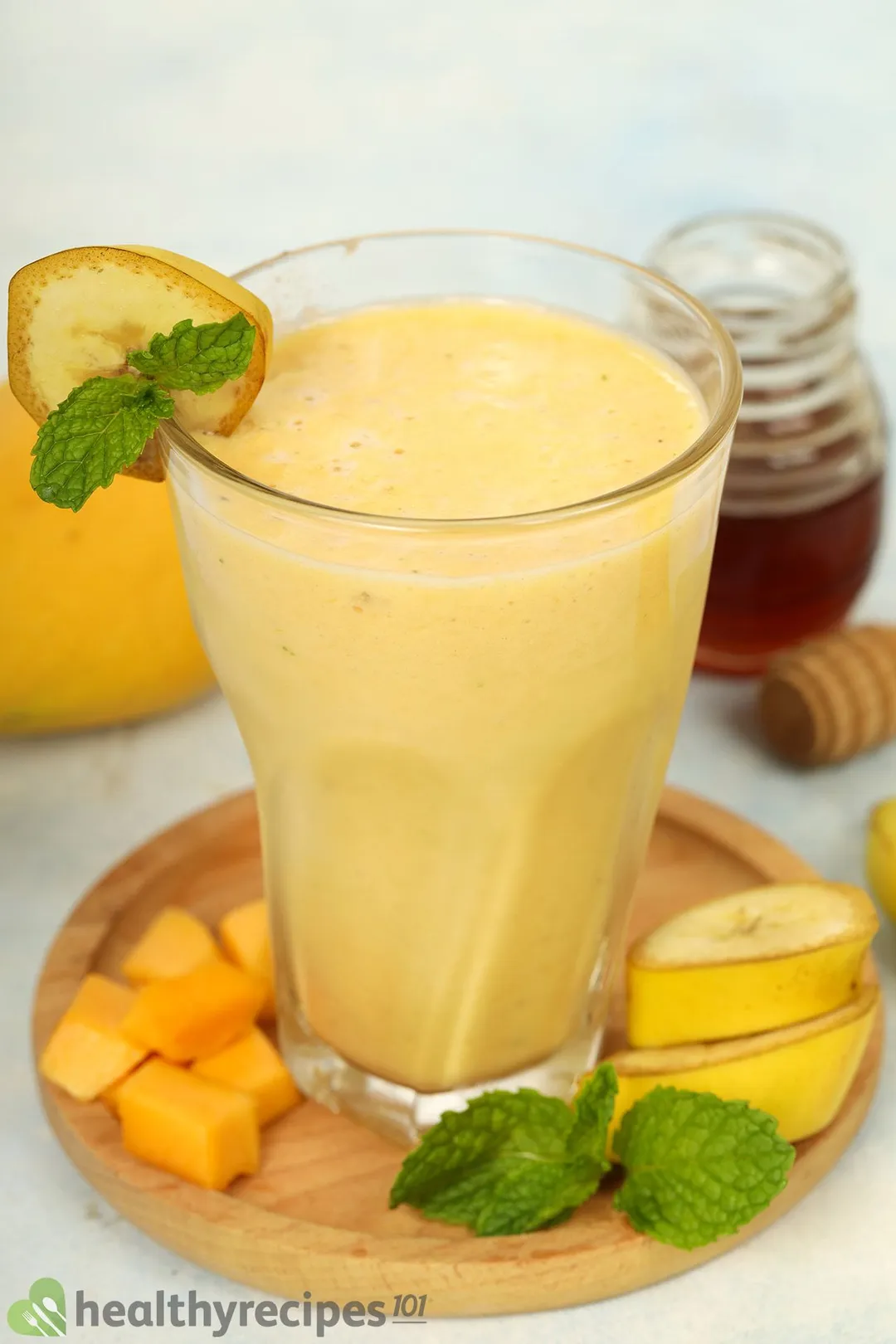 A glass of Mango Banana Smoothie placed on a wooden board with a scored mango, some bananas, and mint leaves.