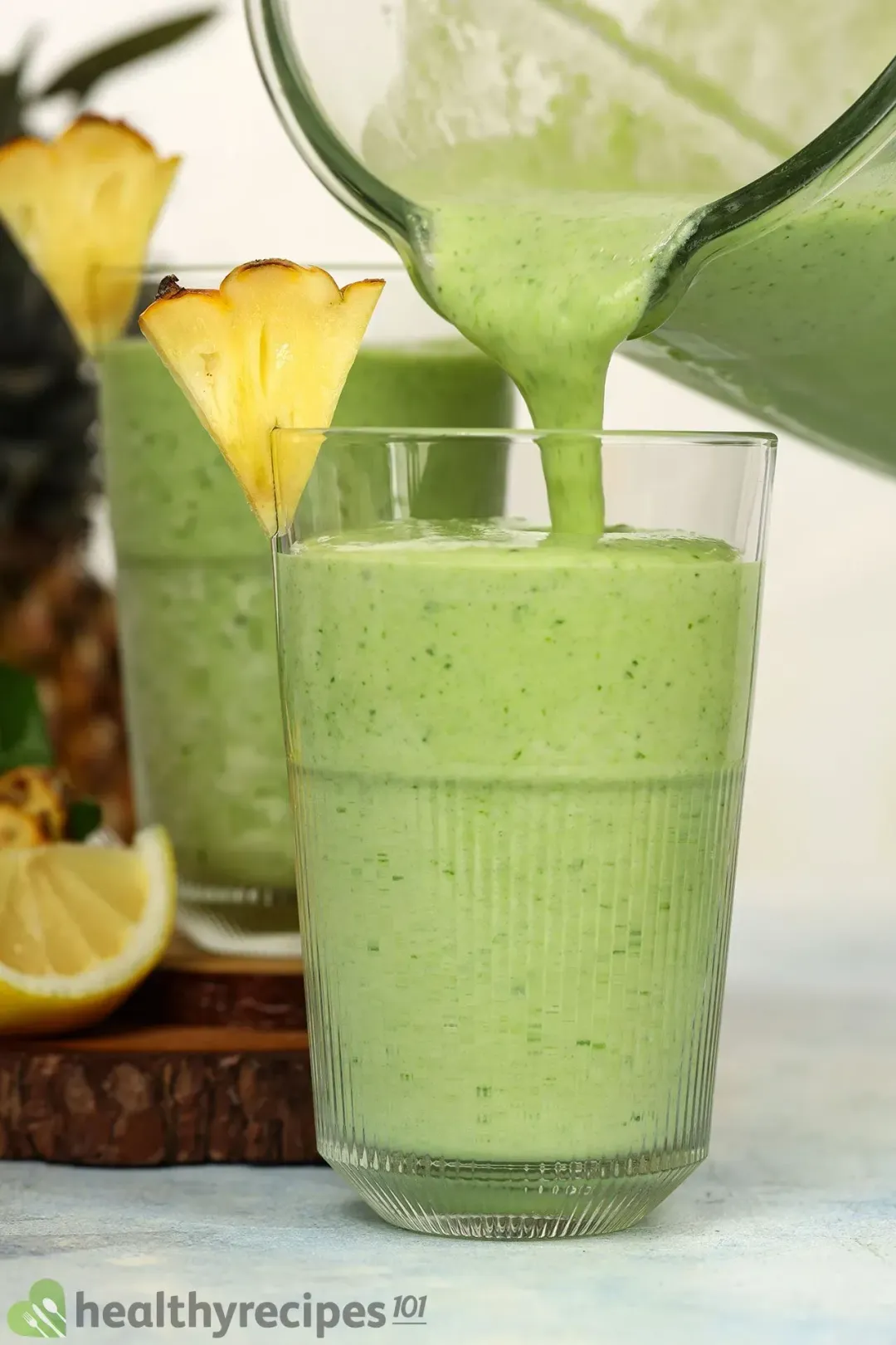 From a pitcher, a green smoothie poured into a glass, garnished with a triangular pineapple wedge