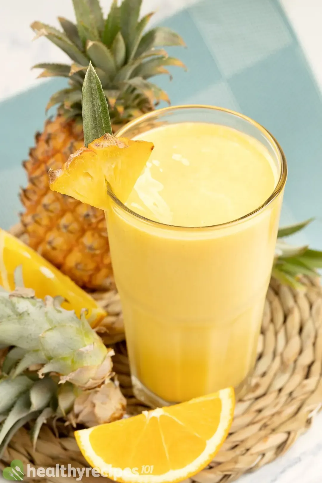 a glass of mango pineapple smoothie next to an orange wedge, a pineapple head, and a whole pineapple