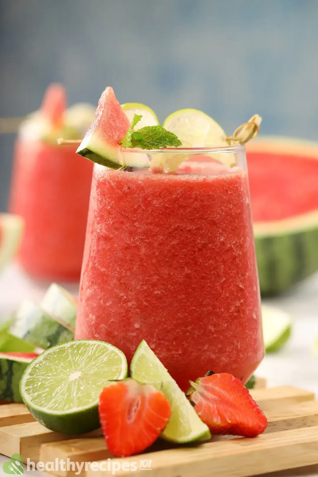A glass of watermelon mojito smoothie, slices of watermelon, halved lime, and strawberries placed on a wooden tray