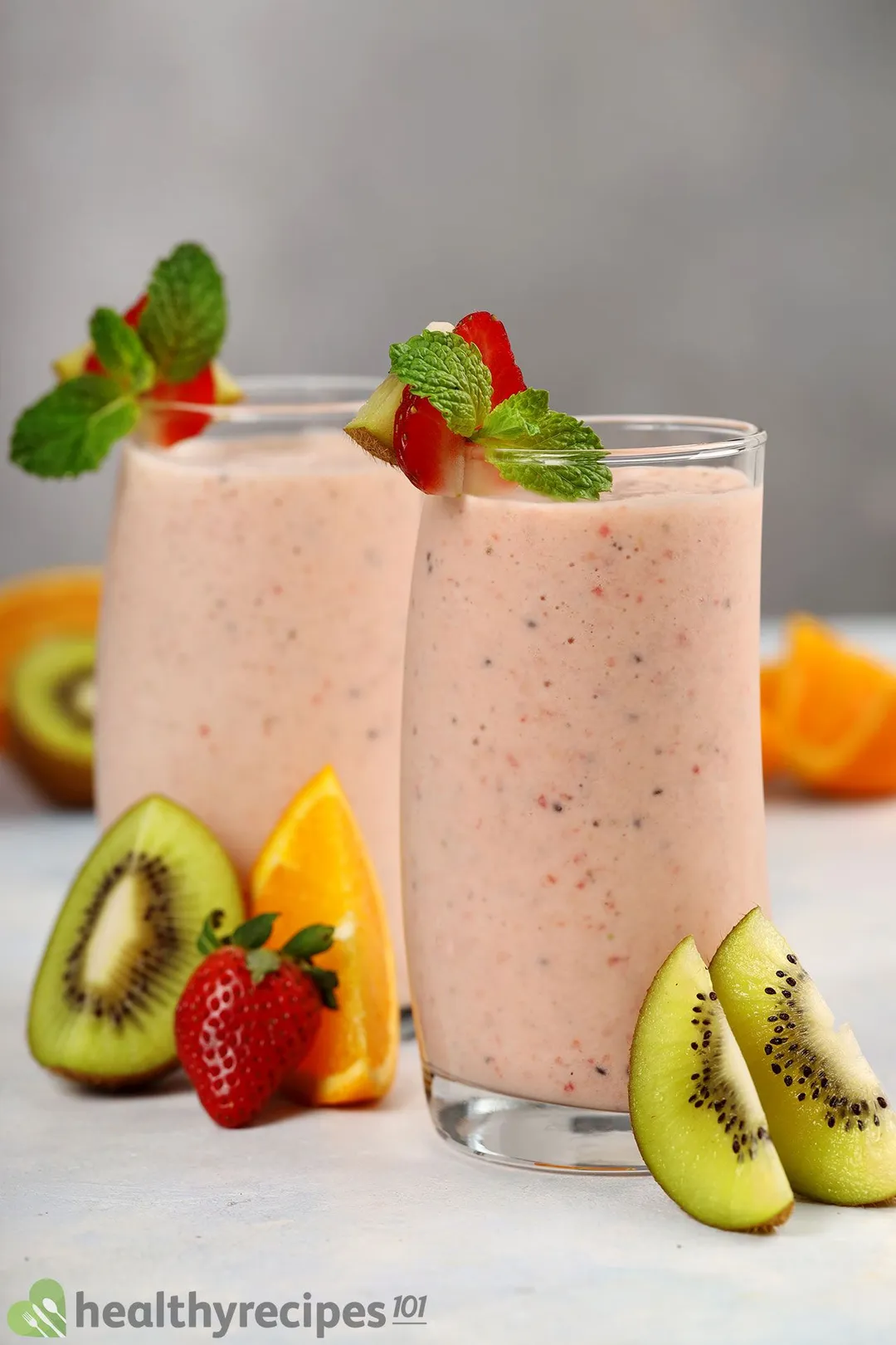 Two glasses of Kiwi Quencher Tropical Smoothie placed near slices of green kiwi, orange, and strawberry.
