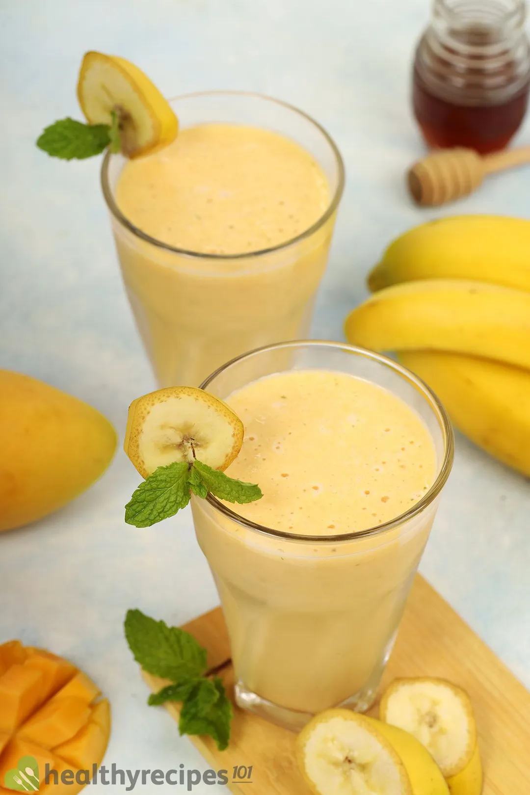 Two glasses of Mango Banana Smoothie placed near mangoes, bananas, and a small jar of honey.
