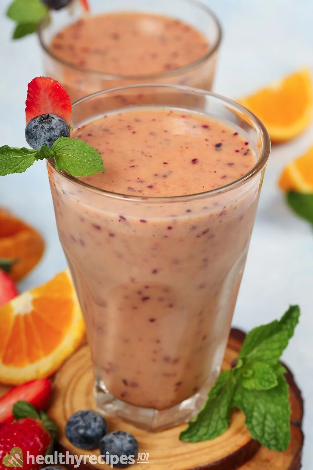 Two glasses of Mango Berry Smoothie placed on a wooden board near strawberries, blueberries, and half an orange.