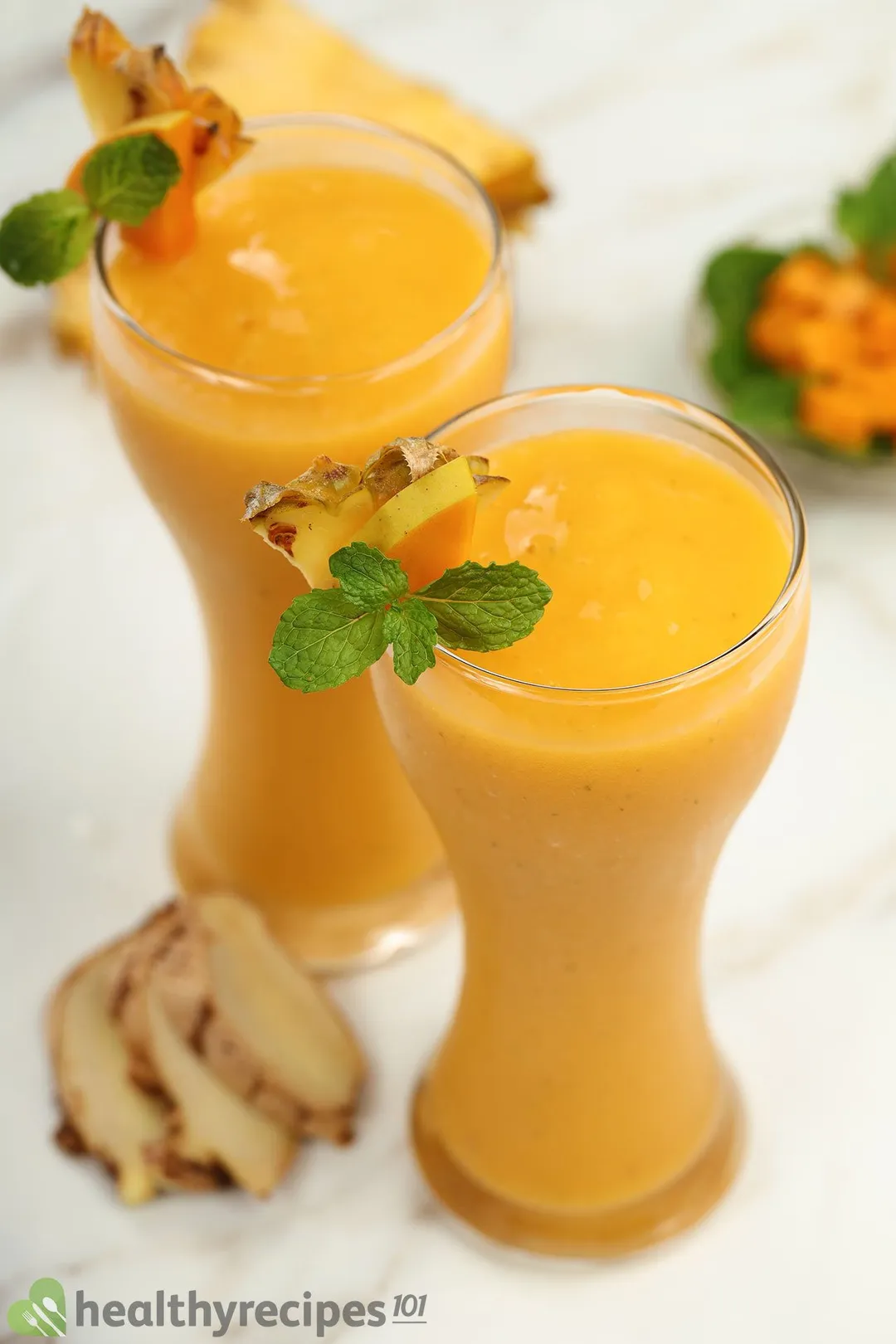 Two tall glasses of Mango Ginger Smoothie decorated with slices of pineapples and mint leaves.
