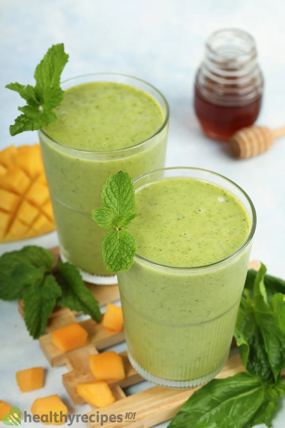 Two glasses of Mango Spinach Smoothie placed on a wooden board near fresh spinach and cubed mangoes.