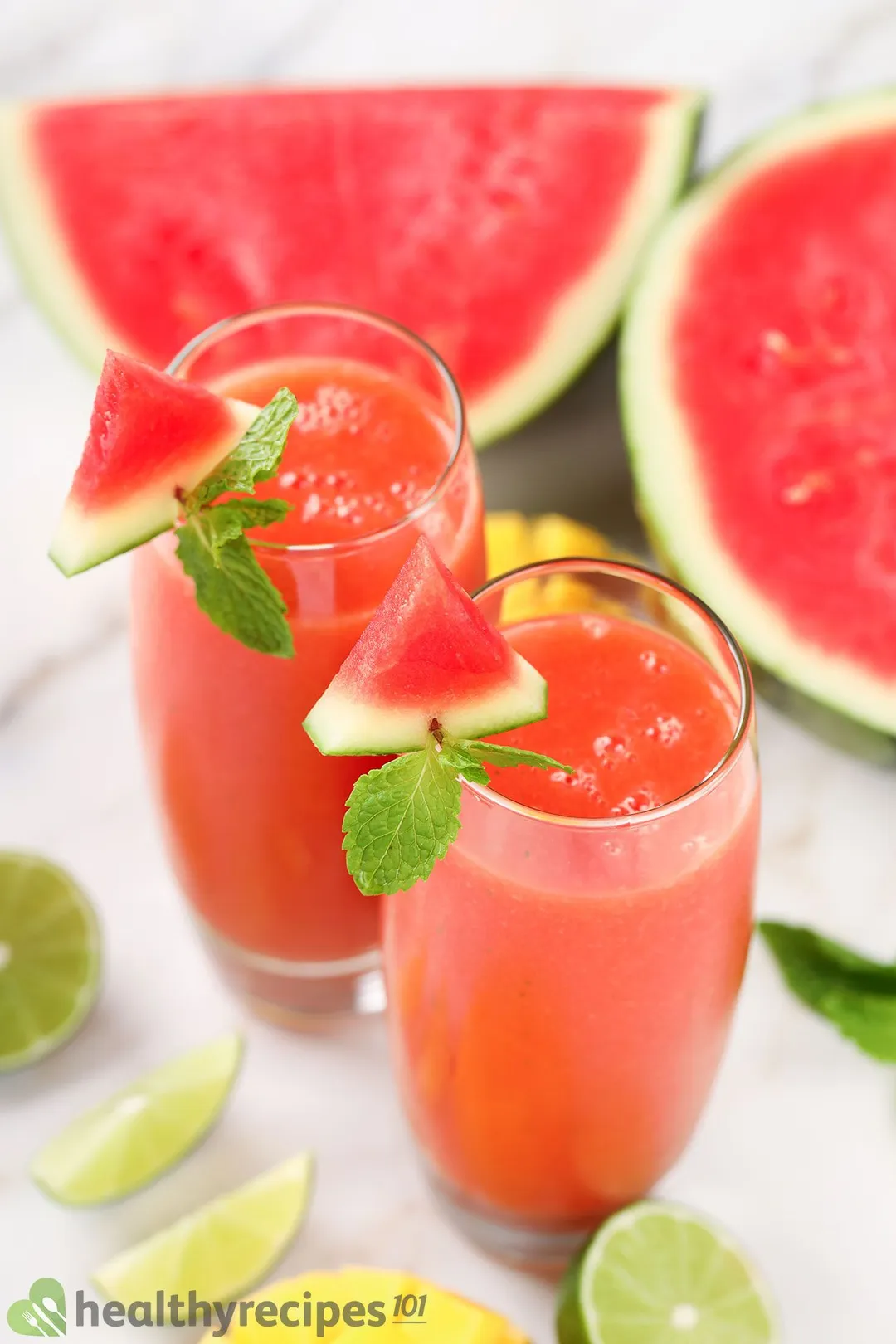 Two glasses of Mango Watermelon Smoothie placed near two halves of a watermelon, lime wedges, and mint leaves.