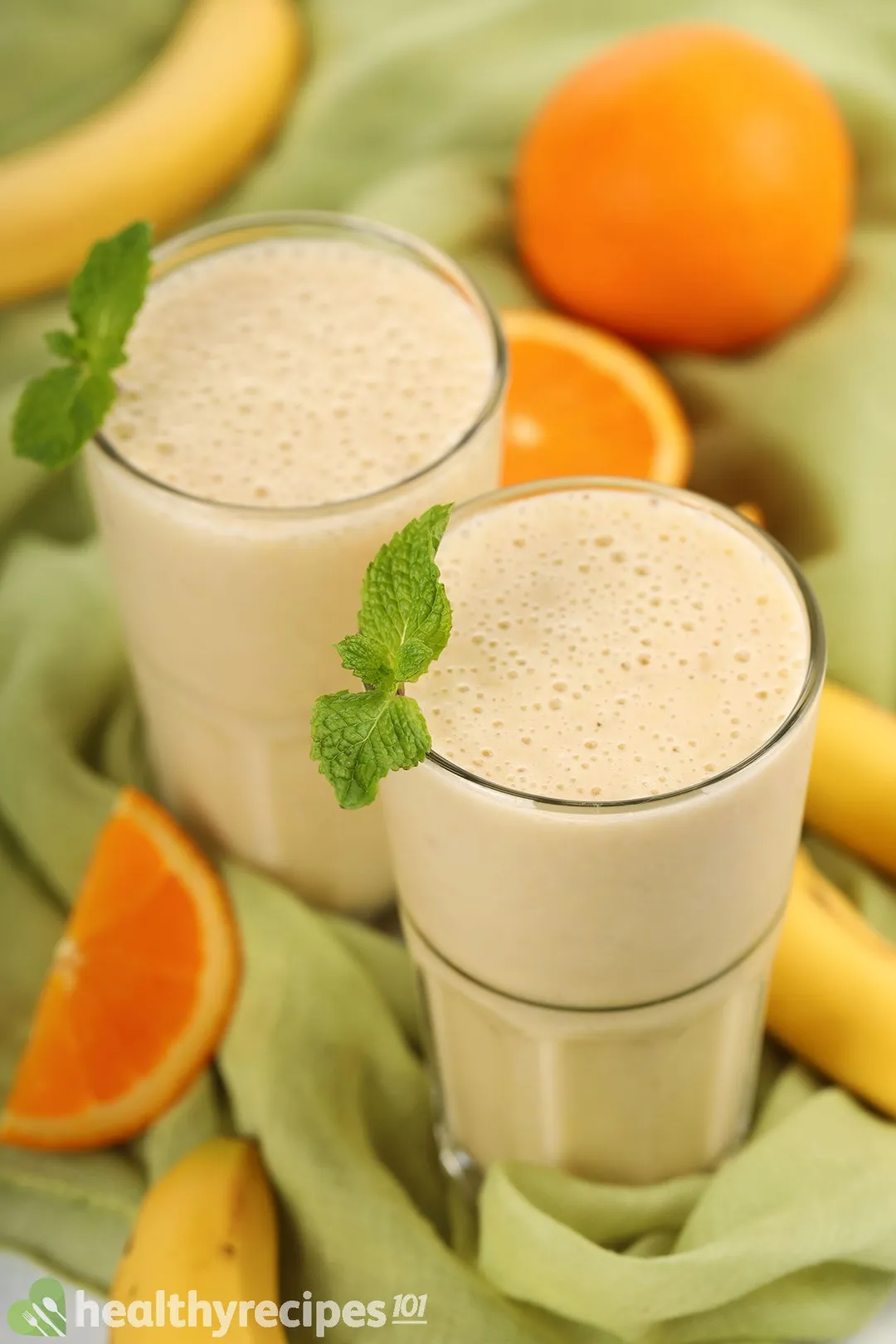 Two glasses of Orange Banana Smoothie placed on a green cloth near orange slices and unpeeled yellow bananas.