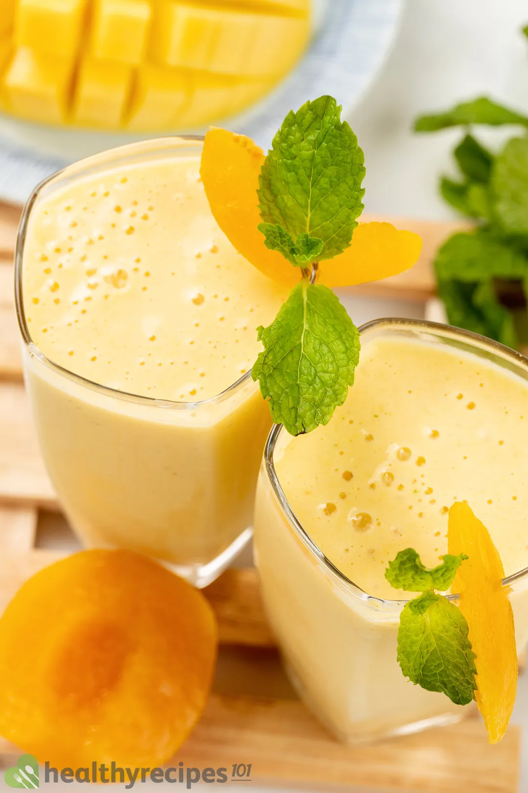 A high-angle shot of two peach smoothie glasses placed on a wooden board and near a sliced peach.