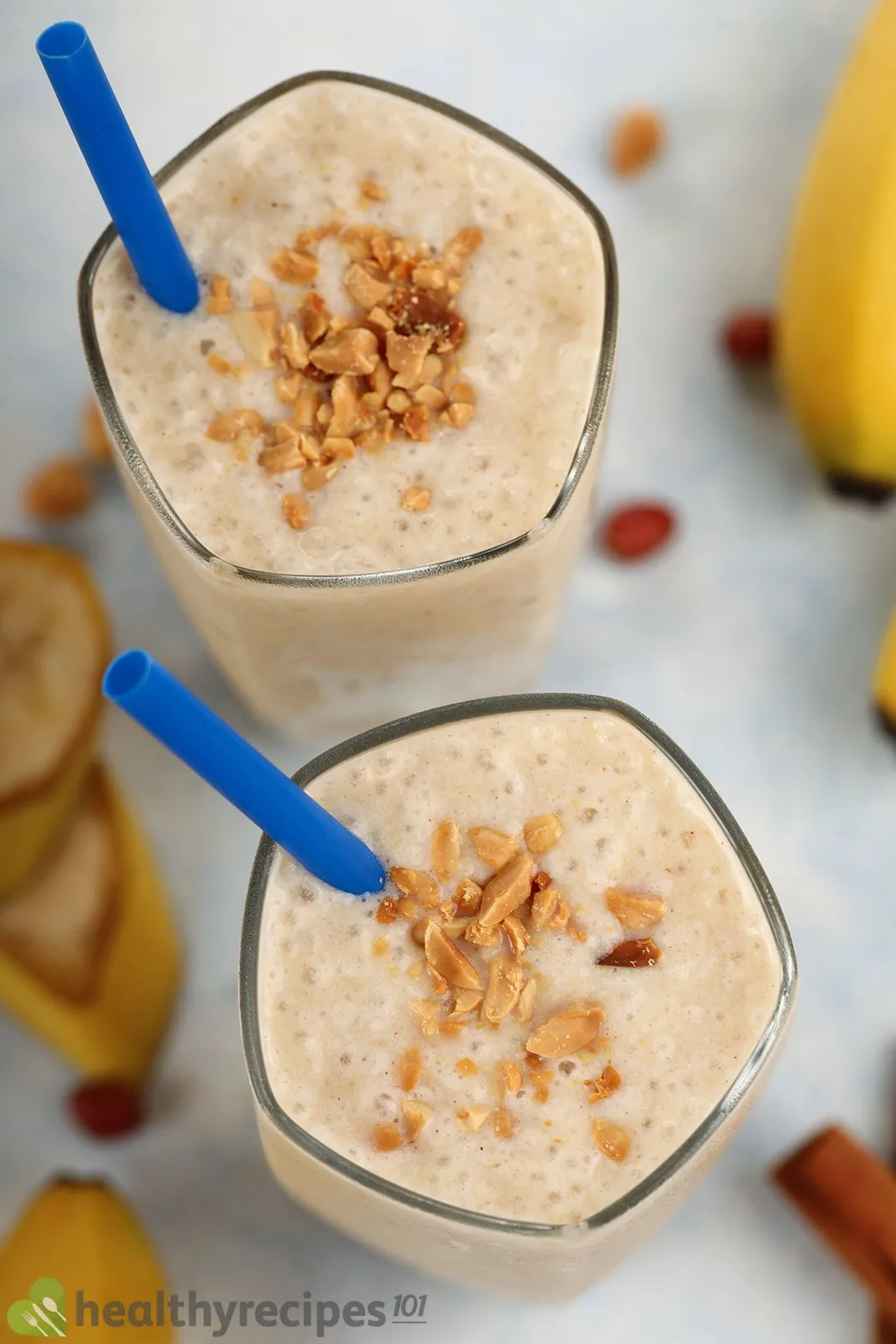 A high angle shot of two glasses of Peanut Butter Banana Smoothie, surrounded by bananas and roasted peanuts.