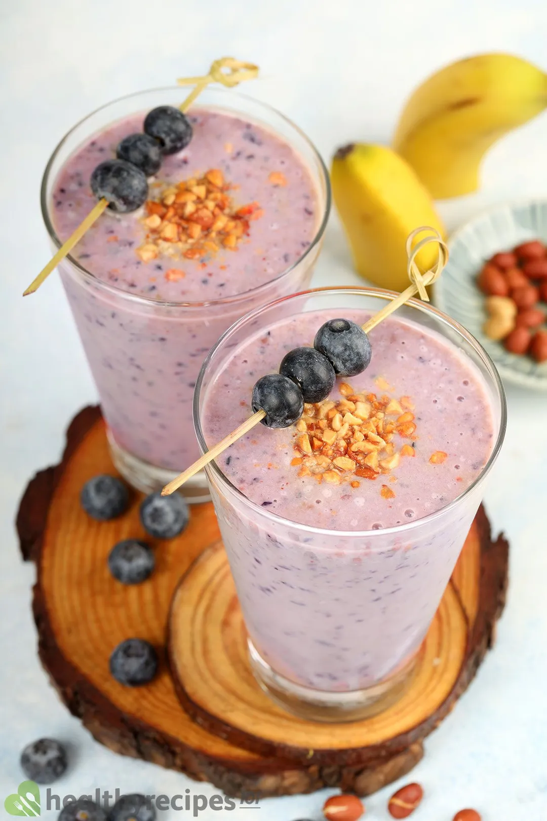 Two glasses of purple peanut butter blueberry smoothie on wooden coasters with some blueberries