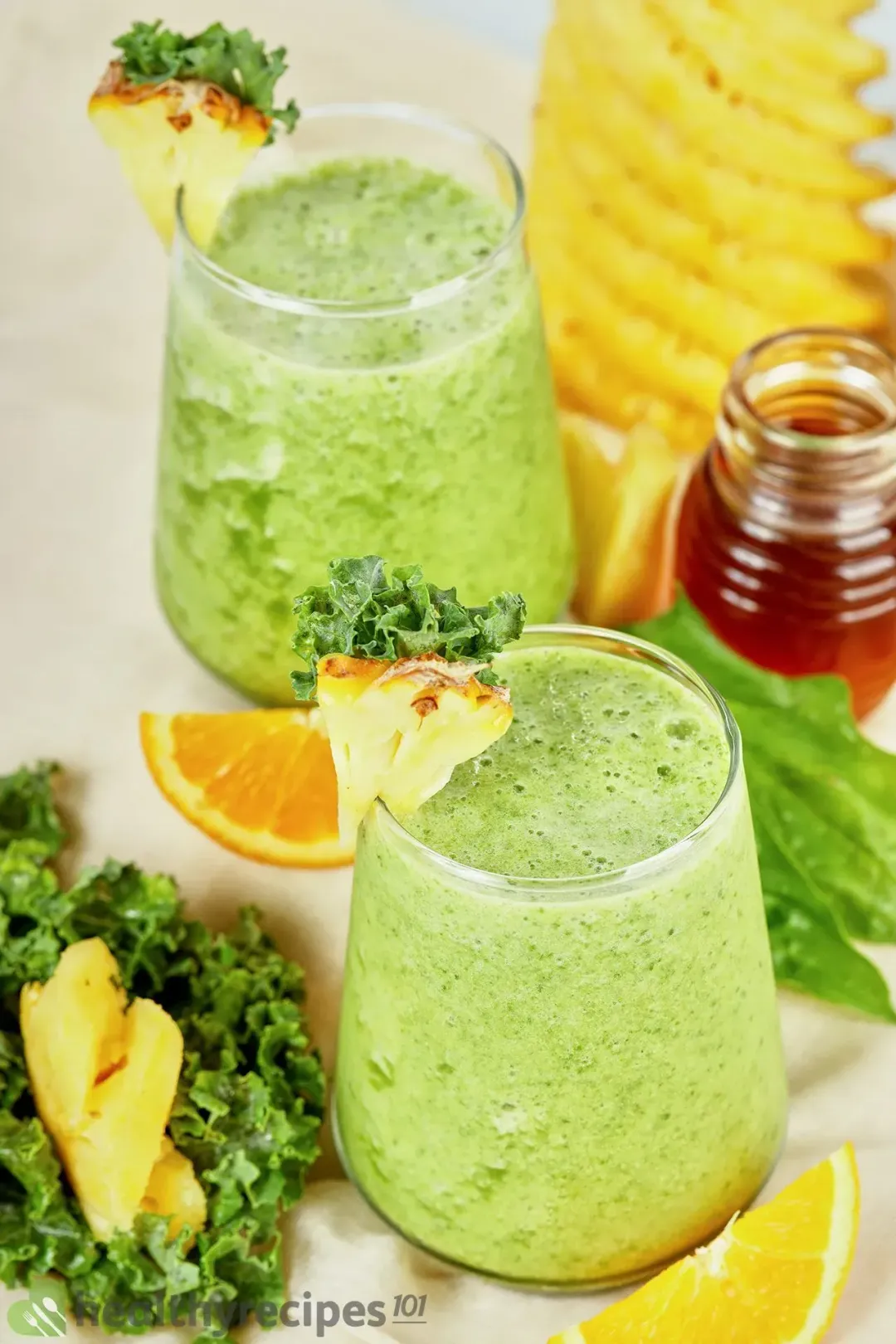 two glasses of pineapple green smoothie with pineapple wedges placed on the rims and surrounded by kale, orange wedges, and other decorants