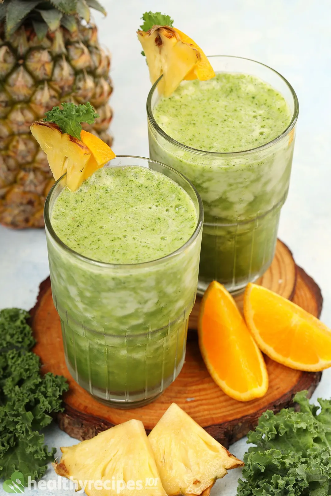 Two glasses of Pineapple Kale Smoothie placed on a wooden board near slices of orange, kale leaves, and a pineapple.