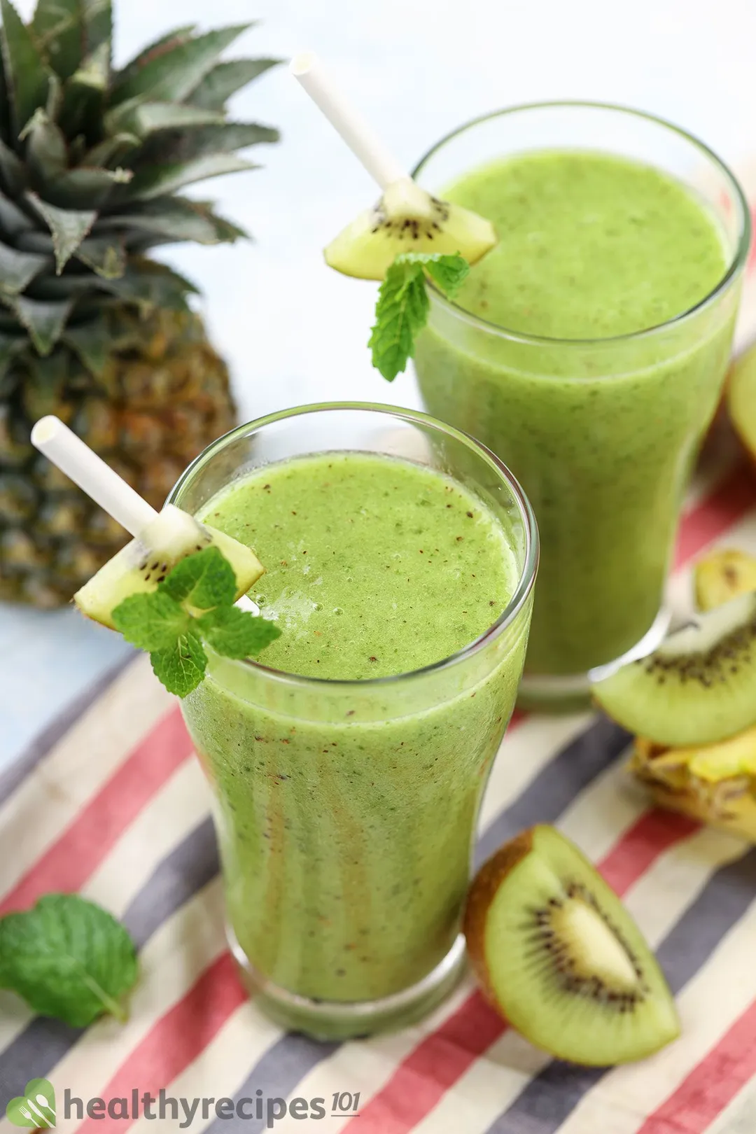 Two glasses of Pineapple Kiwi Smoothie placed on a tablecloth and near slices of kiwi and pineapple.