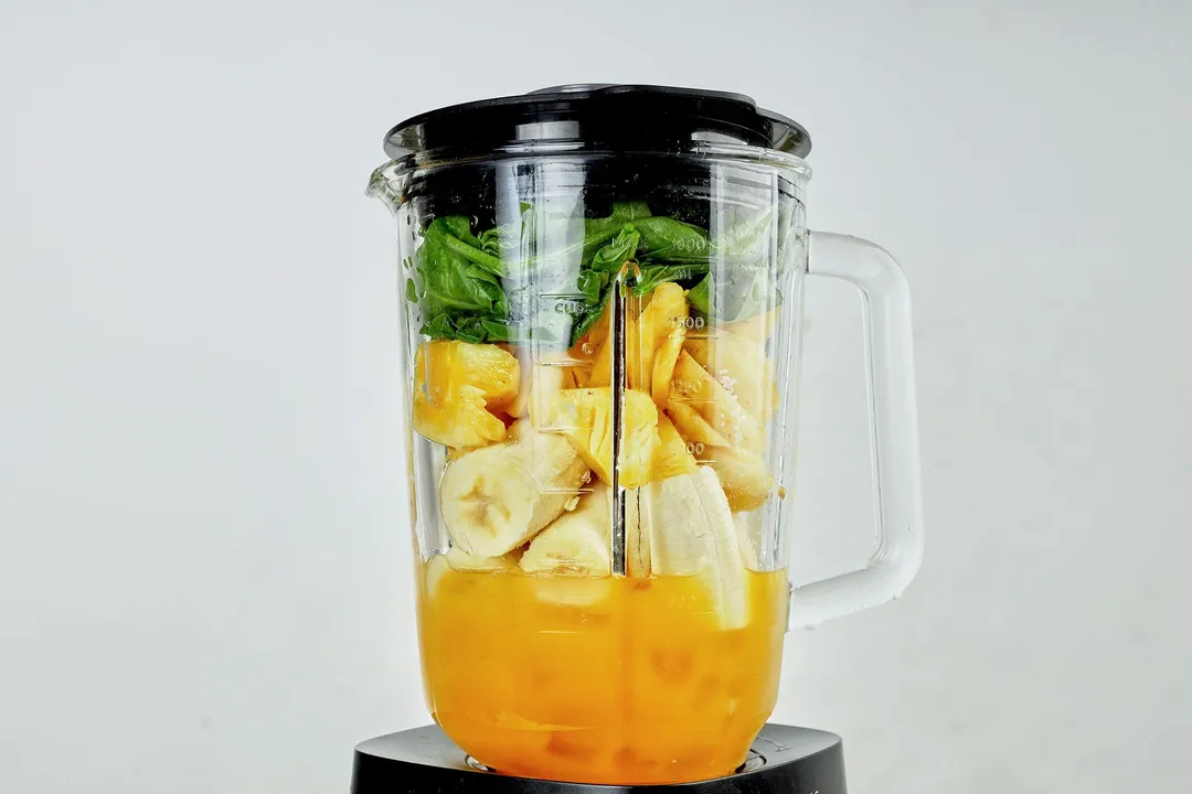 a front shot of a blender pitcher with spinach, cubed banana, cubed pineapple and orange juice in it
