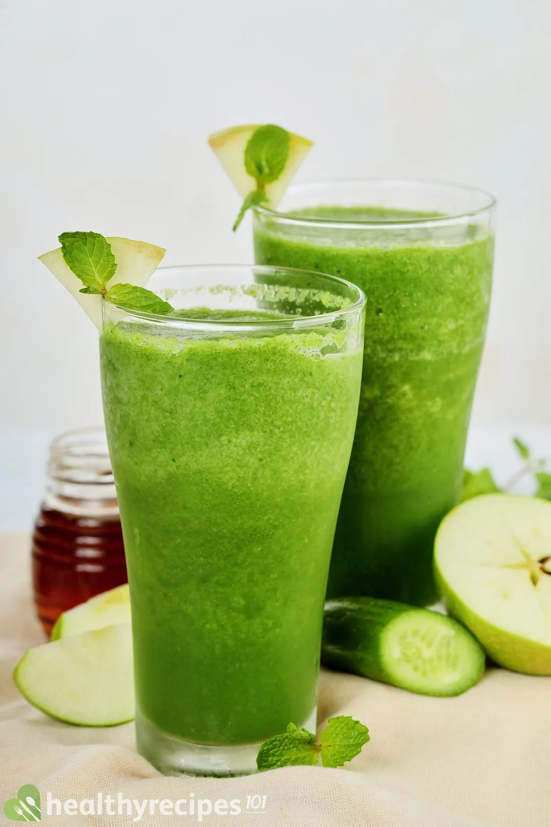 Two tall glasses of sour apple smoothie laid near slices of green apples, cucumbers, and a small jar of honey.