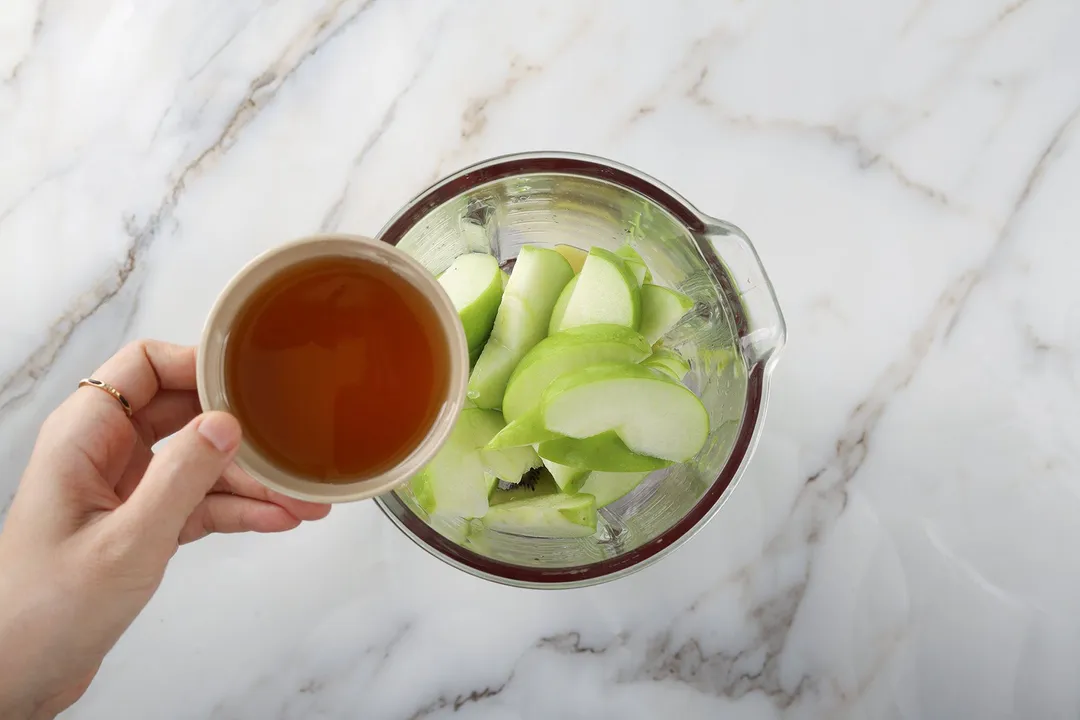 a hand holding a small bowl of honey pouring into a blender of sliced green apple and kiwi