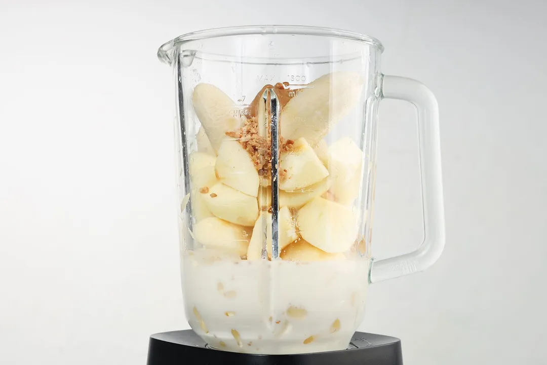 front shot of a blender pitcher with banana, apple cubed, peanut butter, milk and ice in it