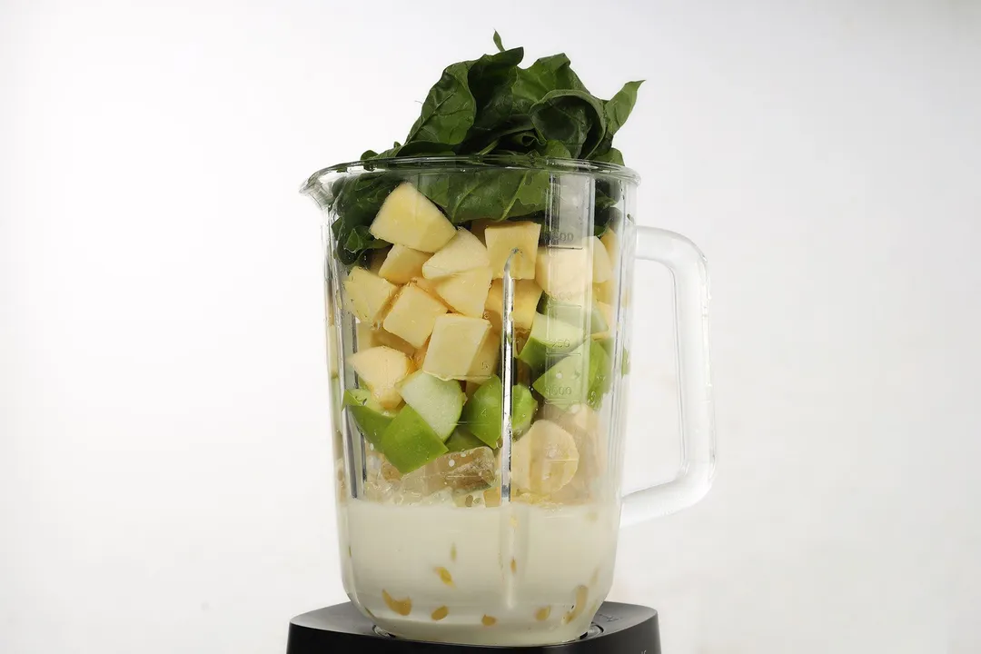 a blender pitcher full of spinach, apple cubed, banana cubed, milk and ice