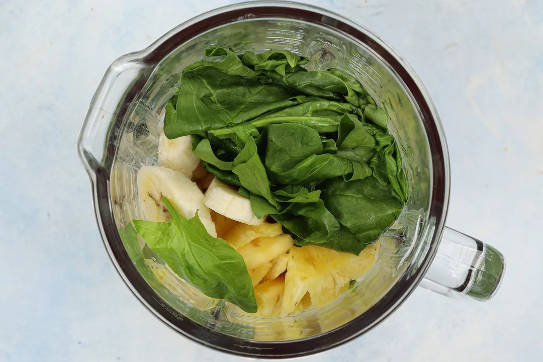 top view of a blender pitcher full of spinach, sliced avocado, sliced banana and pineapple.