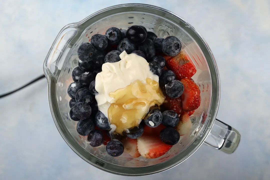 top view of a blender pitcher with honey, yogurt, blueberries and strawberries in it.