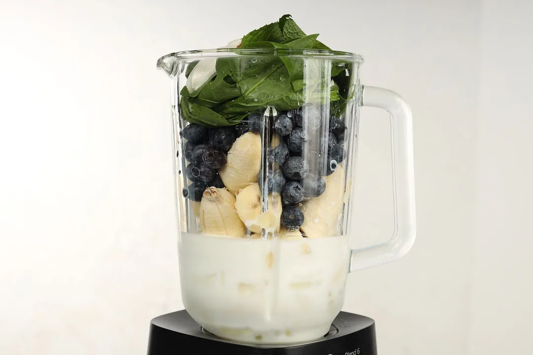front shot of a blender pitcher full of spinach, blueberry, banana, milk and ice
