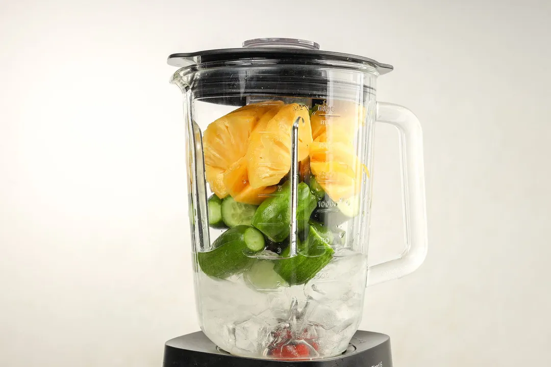 a front shot of a blender pitcher with pineapple cubed, cucumber and ice in it