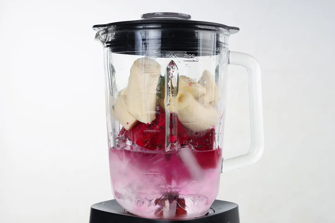 a blender pitcher with banana cubed, dragon fruit cubed and ice in it