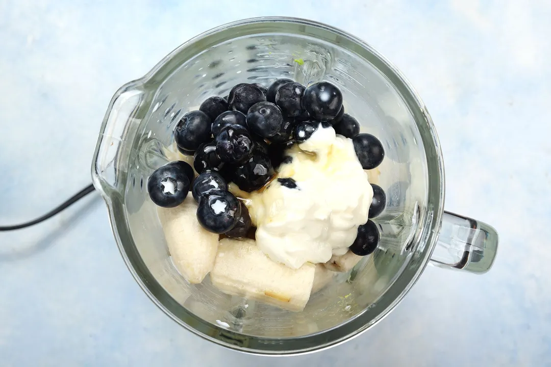 A high-angle shot of a blender filled with blueberries, sliced bananas, and yogurt.