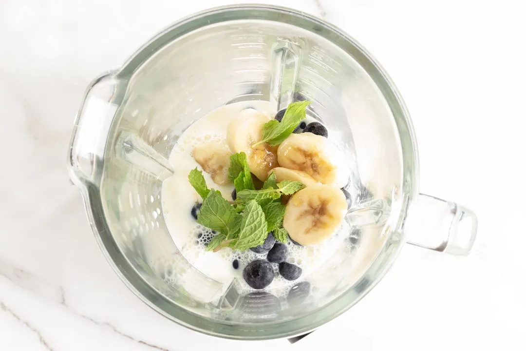 A high-angle shot of a blender filled with sliced bananas, mint leaves, blueberries, and milk.