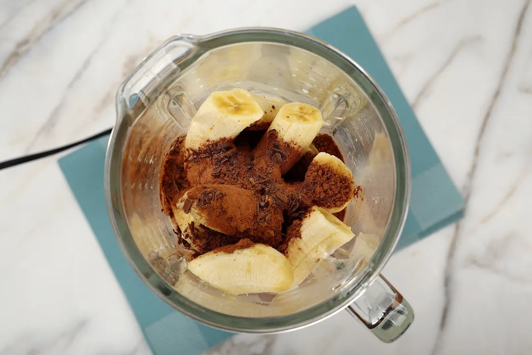 A high-angle shot of a blender filled with sliced peeled bananas, cocoa powder, and minced chocolate.