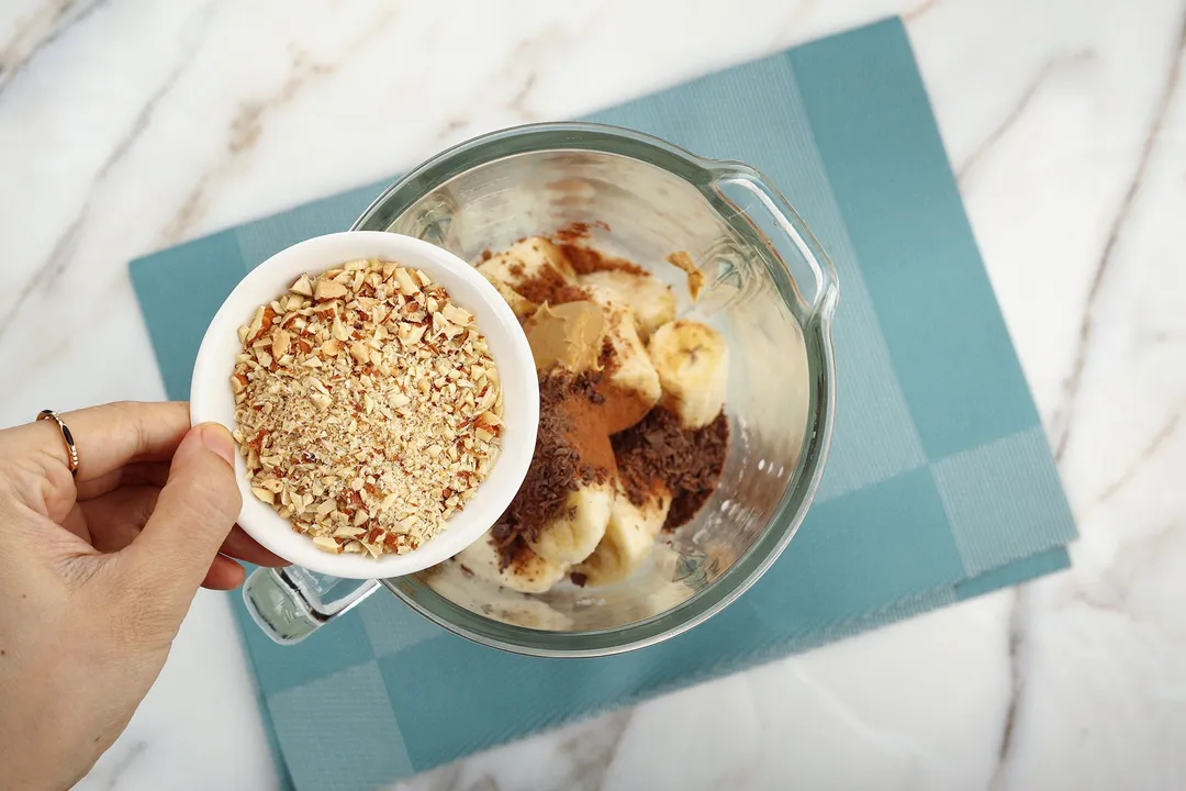 A hand holding a small plate of chopped almonds over a blender filled with sliced and peeled bananas, cocoa powder, chocolate, etc.