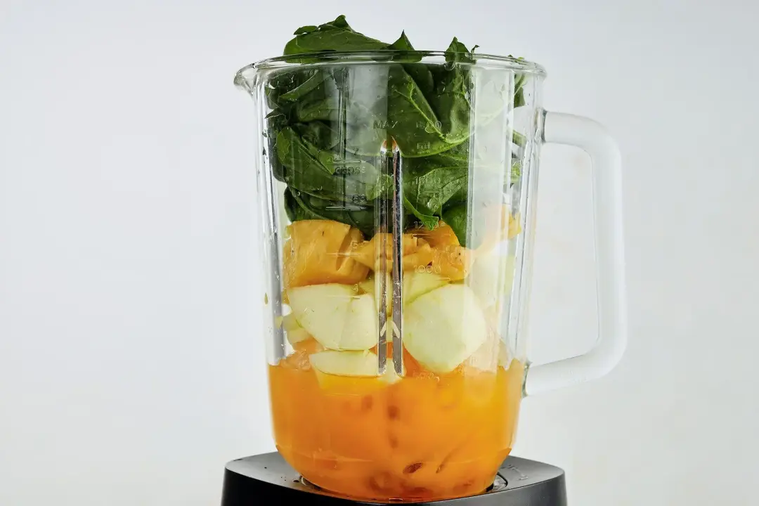 A blender layered with orange juice, large apple cubes, spinach leaves, and ice