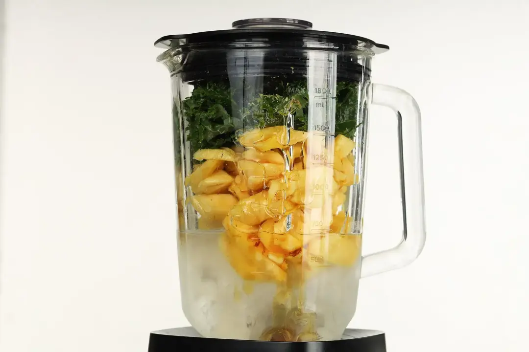 Ingredients of the smoothie packed into a blender in separate layers: coconut water and ice, sliced pineapples, and kale leaves