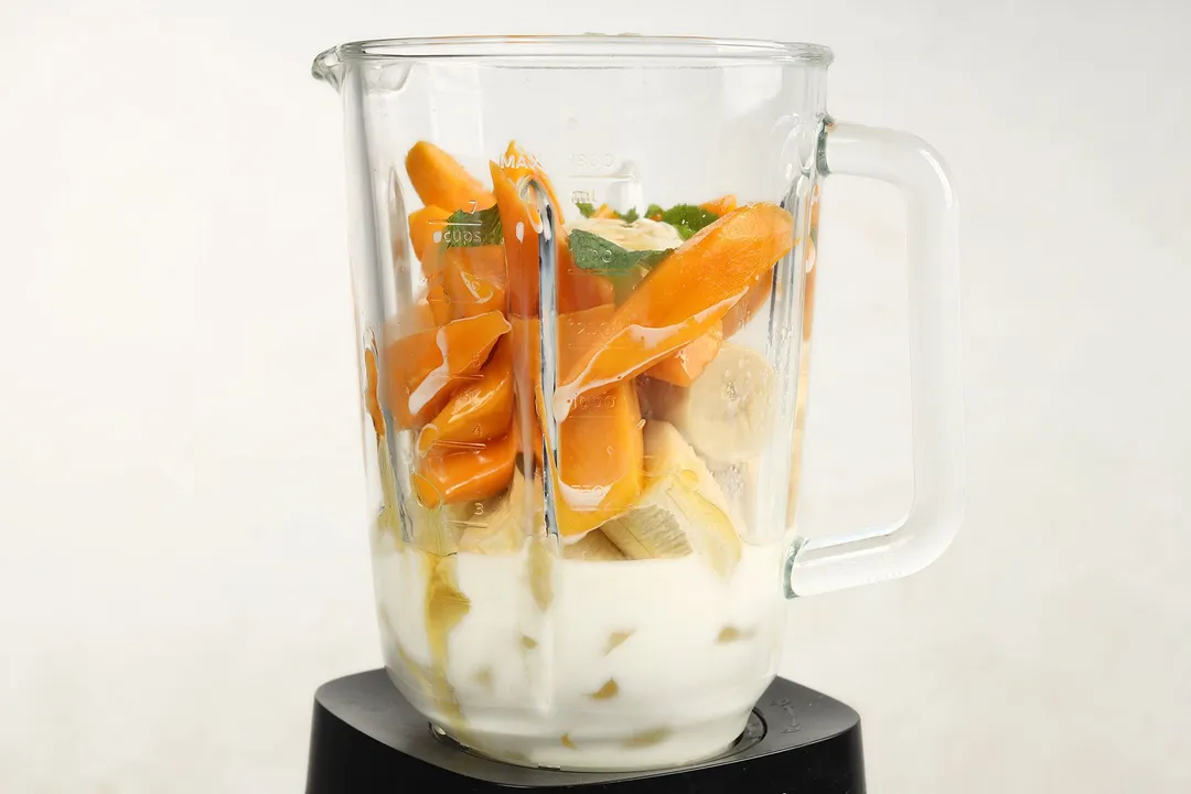 A blender filled with slices of banana, mango, milk, and mint leaves.
