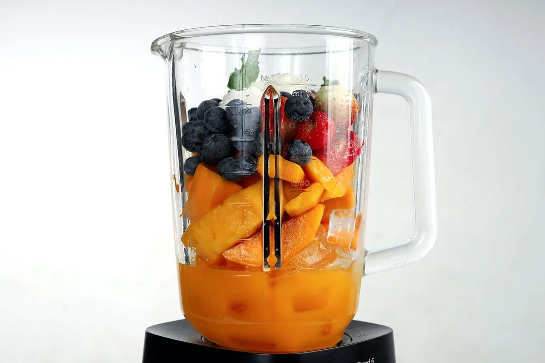 A blender filled with orange juice, mango slices, blueberries, and strawberries.