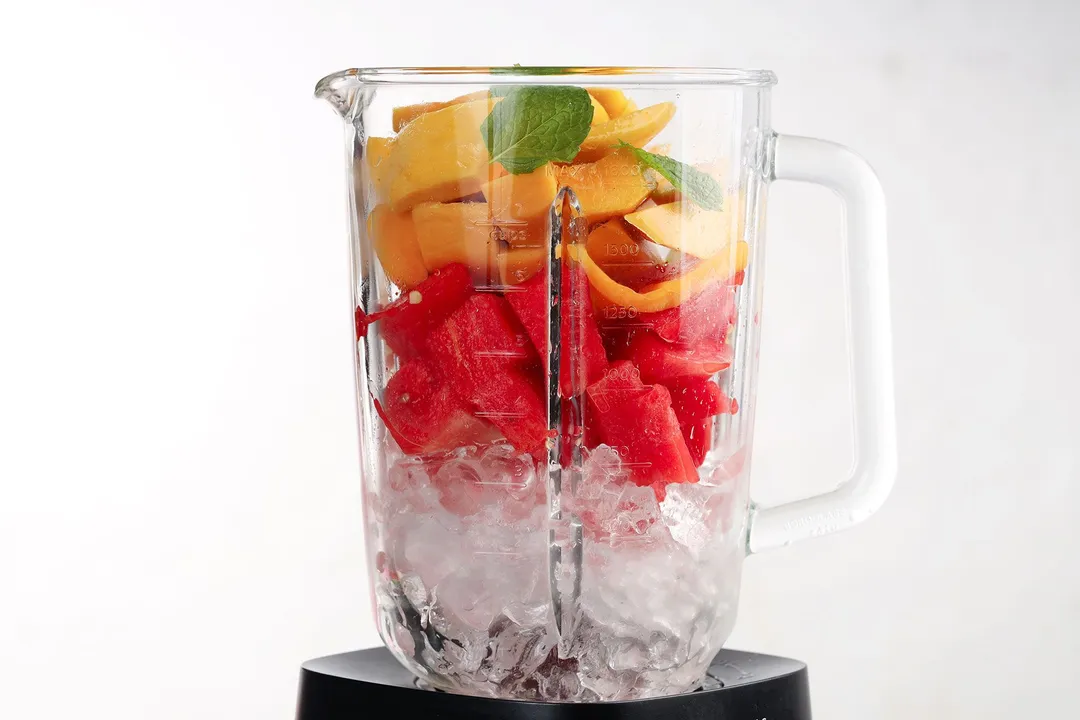A blender filled with ice, watermelon triangles, mango wedges, and mint leaves.