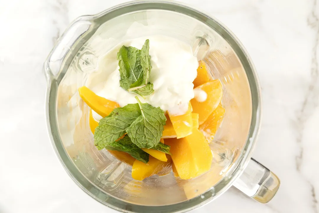 A high-angle shot of a blender filled with peach, mango, mint leaves, and yogurt.