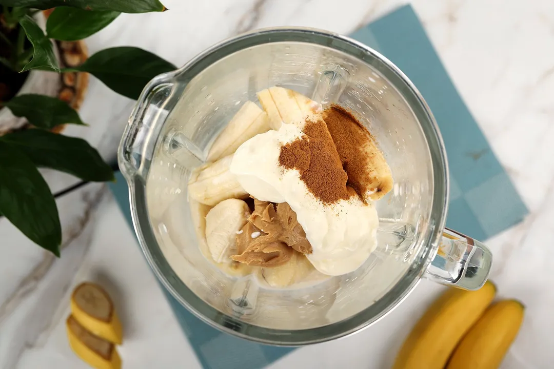 A high-angle shot of a blender filled halfway with peeled banana, cinnamon powder, peanut butter, and yogurt.